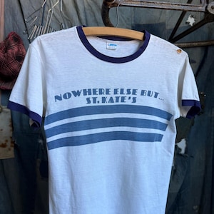 May include: A white ringer t-shirt with dark blue trim on the collar and sleeve cuffs. The front features the blue text "NOWHERE ELSE BUT... ST. KATE'S" above three horizontal blue stripes. A small Champion brand tag is visible at the neckline.