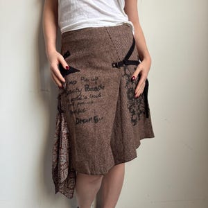 May include: Brown tweed skirt with a unique asymmetrical design. Features black accents, a decorative fabric panel, and handwritten text. The skirt has a wrap-style closure with a buckle.