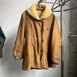 May include: A vintage brown coat with a tan shearling collar. The coat has a double-breasted design with dark buttons and pockets. The sleeves have dark trim. The coat is hanging on a metal rack.