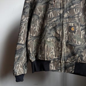 Puede incluir: Chaqueta de camuflaje Carhartt con cierre de cremallera completo. La chaqueta presenta un patr&oacute;n de corteza de &aacute;rbol marr&oacute;n, verde y beige. Tiene cuello y pu&ntilde;os de canal&eacute; negros y un parche con el logo de Carhartt en el bolsillo delantero.