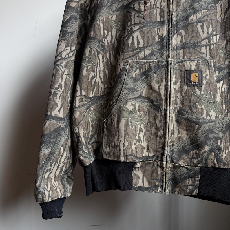 Puede incluir: Chaqueta de camuflaje Carhartt con cierre de cremallera completo. La chaqueta presenta un patr&oacute;n de corteza de &aacute;rbol marr&oacute;n, verde y beige. Tiene cuello y pu&ntilde;os de canal&eacute; negros y un parche con el logo de Carhartt en el bolsillo delantero.