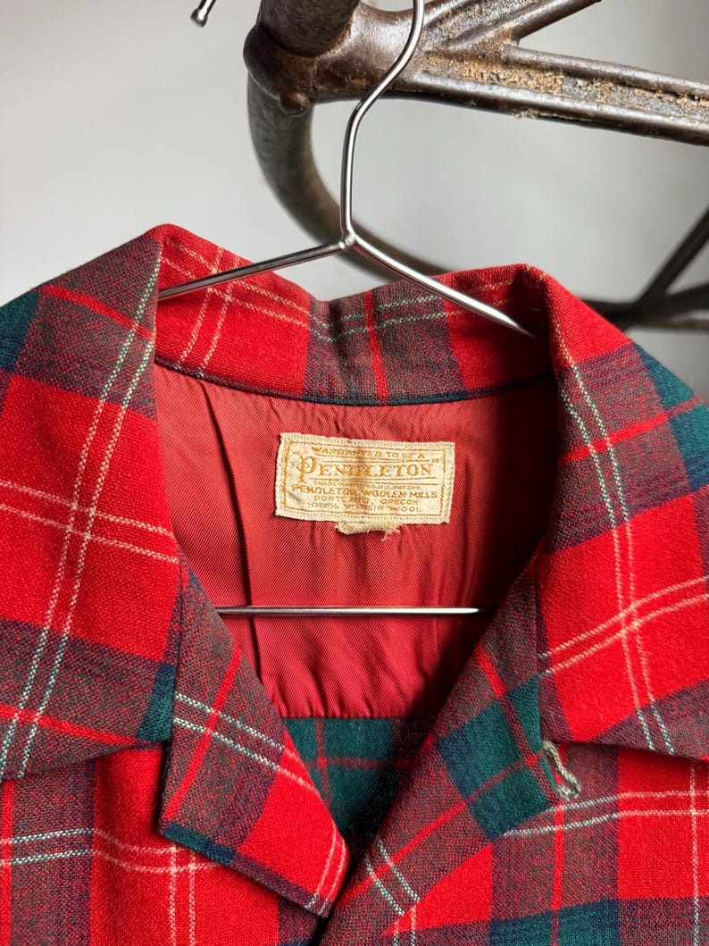 K&ouml;nnte beinhalten: Eine rot, gr&uuml;n und wei&szlig; karierte Jacke mit Kragen. Die Jacke h&auml;ngt an einem Metallb&uuml;gel. Das Etikett in der Jacke lautet "Pendleton" und enth&auml;lt Text.
