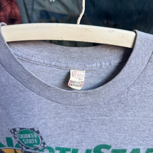 May include: A heather gray t-shirt with a ribbed crew neckline. A white fabric tag inside the collar displays the size L and the Screen Stars brand logo in red. A partial graphic featuring a green checkered flag with the text Quaker State is visible.
