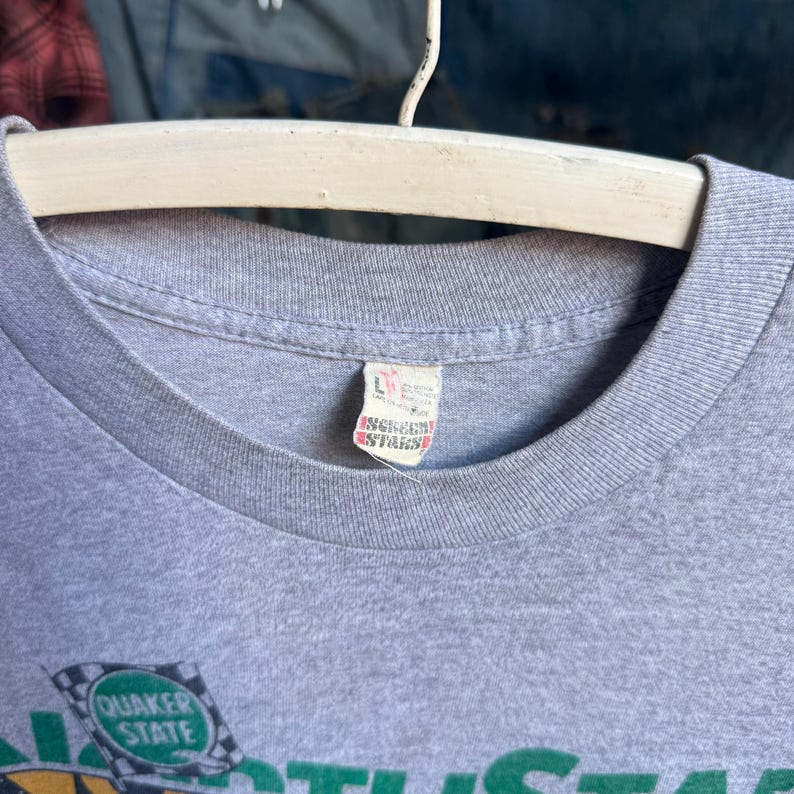 May include: A heather gray t-shirt with a ribbed crew neckline. A white fabric tag inside the collar displays the size L and the Screen Stars brand logo in red. A partial graphic featuring a green checkered flag with the text Quaker State is visible.