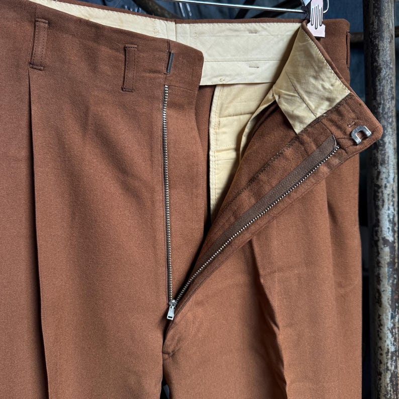 May include: Brown trousers featuring a high-waisted design with a metal zipper fly, belt loops, and a light beige interior lining. The garment includes a single pleat on the front and a metal hook-and-bar closure at the waistband.