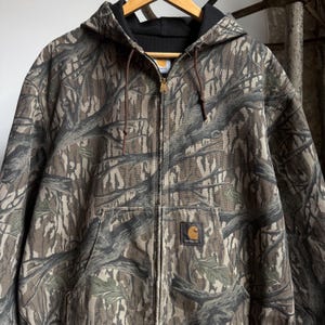 Puede incluir: Chaqueta con capucha Carhartt con cierre de cremallera completo y estampado de camuflaje. La chaqueta tiene un cord&oacute;n marr&oacute;n, un cuello negro y un bolsillo delantero con el logotipo de Carhartt. El dise&ntilde;o de camuflaje incorpora tonos marrones, verdes y grises.