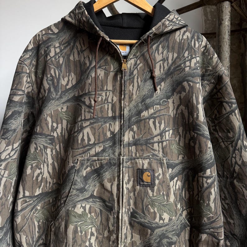 Puede incluir: Chaqueta con capucha Carhartt con cierre de cremallera completo y estampado de camuflaje. La chaqueta tiene un cord&oacute;n marr&oacute;n, un cuello negro y un bolsillo delantero con el logotipo de Carhartt. El dise&ntilde;o de camuflaje incorpora tonos marrones, verdes y grises.