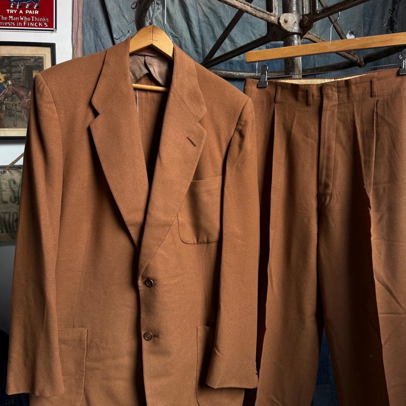May include: A solid brown two-piece suit consisting of a single-breasted blazer with notched lapels, two front buttons, and a chest pocket, paired with matching pleated trousers featuring a high waistband and belt loops.