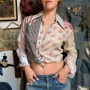 May include: A long-sleeve button-down shirt featuring a patchwork design of various fabrics, including vertical black and white stripes, cream fabric with pink floral patterns, and light blue paisley accents, tied at the waist with blue denim jeans.