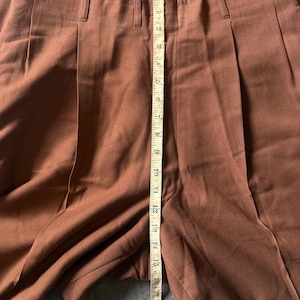 May include: Solid brown trousers featuring a high-waisted design with vertical pleats and belt loops. A yellow measuring tape is placed vertically along the center, showing a length measurement of approximately 19 inches from the waist.