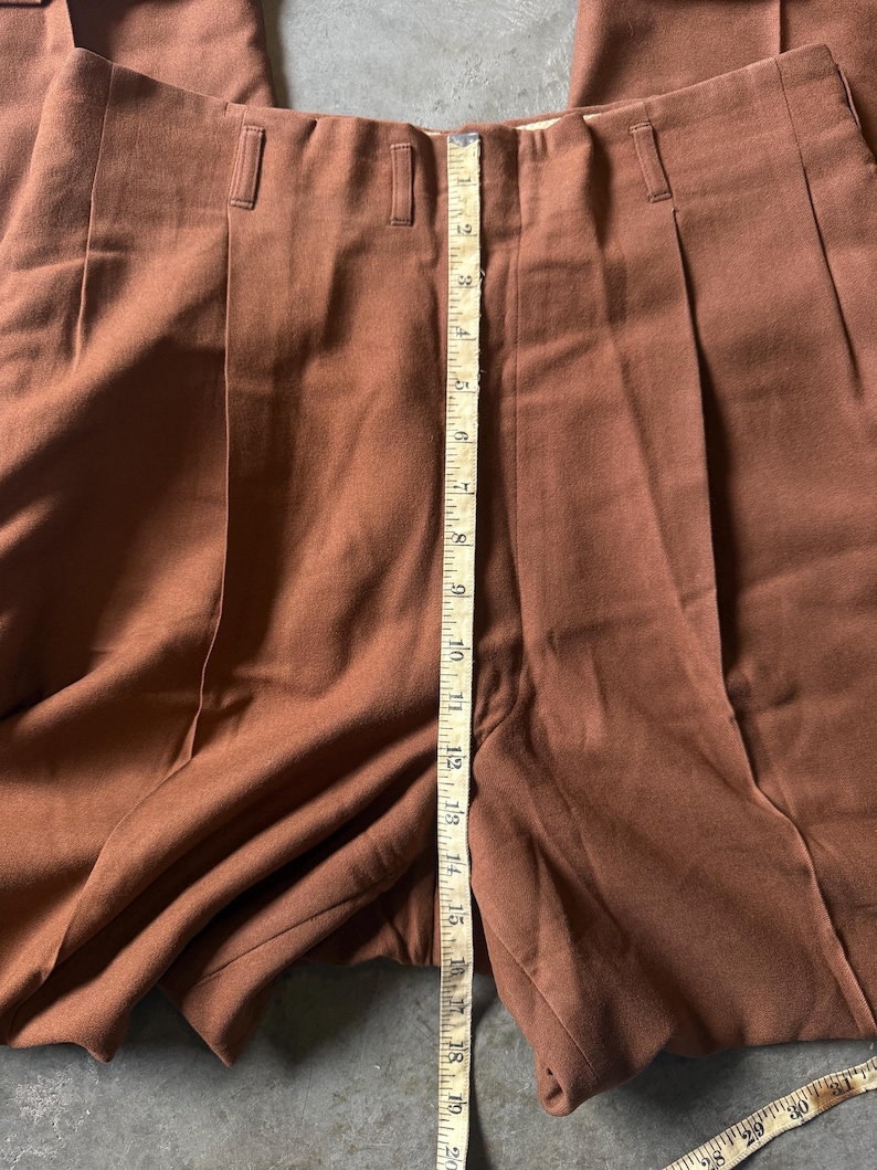 May include: Solid brown trousers featuring a high-waisted design with vertical pleats and belt loops. A yellow measuring tape is placed vertically along the center, showing a length measurement of approximately 19 inches from the waist.