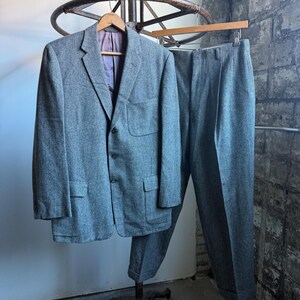 May include: A gray tweed suit, including a blazer and trousers, hangs on a vintage metal clothing rack. The blazer has two front pockets and a purple lining. The trousers are neatly pressed. The suit is a classic style.