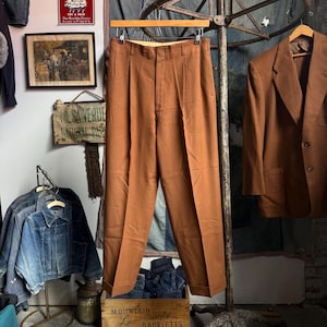 May include: A pair of solid rust-colored trousers featuring a high-waisted design with double front pleats, a button and zipper fly closure, and cuffed hems. The trousers are displayed on a wooden hanger against a neutral background.
