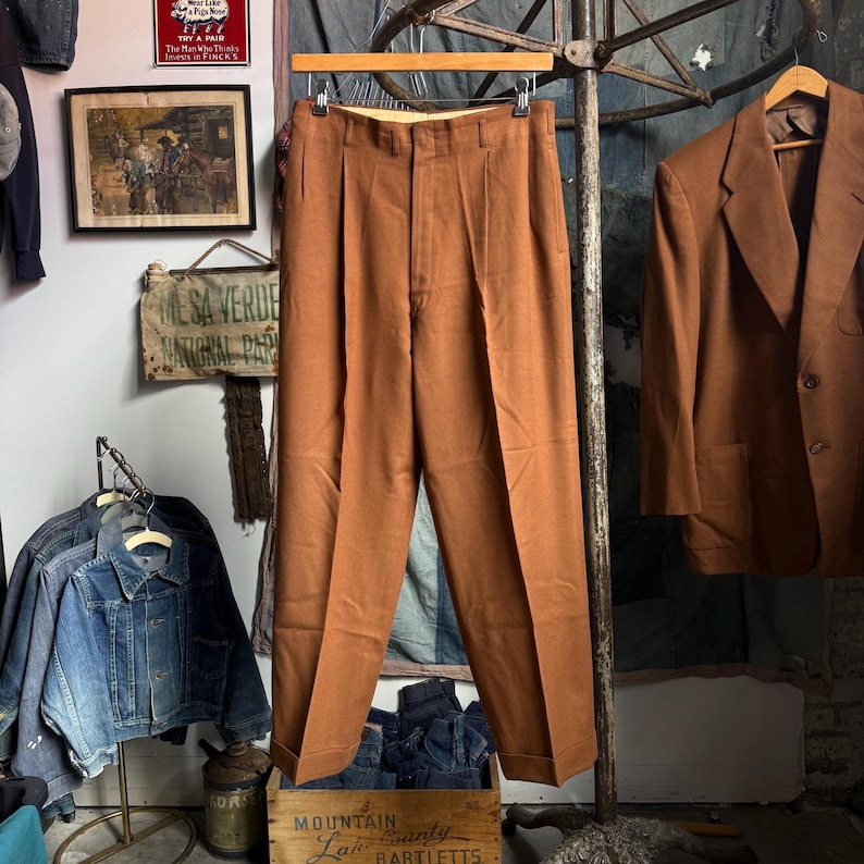 May include: A pair of solid rust-colored trousers featuring a high-waisted design with double front pleats, a button and zipper fly closure, and cuffed hems. The trousers are displayed on a wooden hanger against a neutral background.