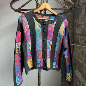 May include: Black cardigan sweater with colorful geometric patterns in pink, blue, yellow, and green. The sweater has a round neckline, long sleeves, and a button-down front. The sweater is hanging on a wooden hanger.