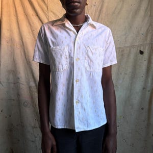 May include: A short-sleeved button-down shirt in white featuring a repeating pattern of small, light brown diamond shapes. The shirt includes a classic collar, a front button placket, and two chest pockets, one on each side of the front panel.