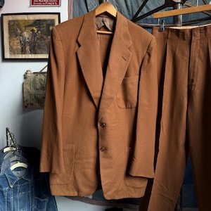 May include: A solid brown suit consisting of a single-breasted blazer and matching trousers. The blazer features notched lapels, two front buttons, a chest pocket, and two lower patch pockets. The trousers have a pleated front and a straight leg.