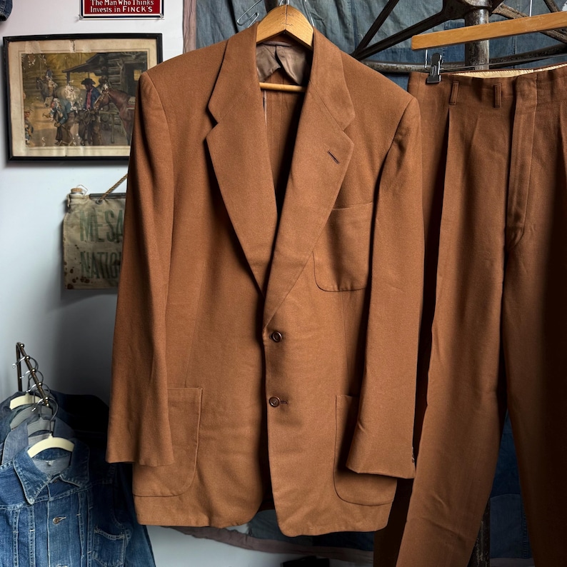 May include: A solid brown suit consisting of a single-breasted blazer and matching trousers. The blazer features notched lapels, two front buttons, a chest pocket, and two lower patch pockets. The trousers have a pleated front and a straight leg.