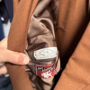 May include: A brown coat with a smooth brown interior lining. The interior features a white rectangular label with the text Amalgamated Clothing Workers of America and a red rectangular label with the text Hart Schaffner & Marx and a white horse logo.