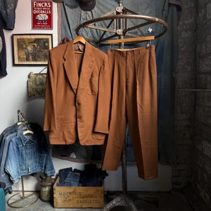 May include: A solid brown two-piece suit consisting of a single-breasted blazer with two front buttons and two patch pockets, paired with matching pleated trousers. The blazer features notched lapels and a chest pocket, while the trousers have a straight-leg cut.