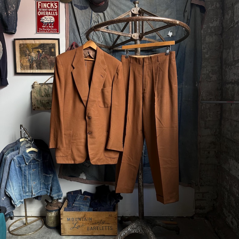 May include: A solid brown suit consisting of a two-button blazer with notched lapels and two front patch pockets, paired with matching pleated trousers. The suit is displayed on a wooden hanger suspended from a metal rack.