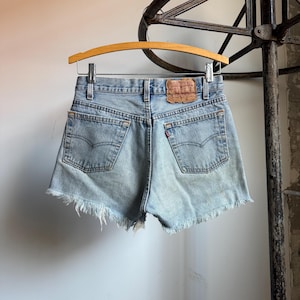 May include: Light blue denim shorts with frayed edges, hanging on a wooden hanger. The shorts feature classic five-pocket styling, a leather patch, and a red Levi's tag. The shorts are vintage and show signs of wear.