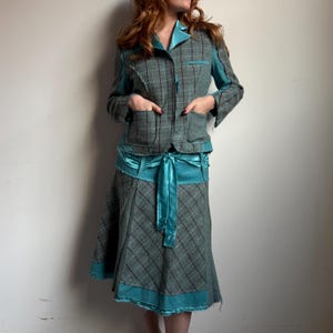 May include: A two-piece teal and gray plaid suit. The jacket has pockets and teal accents on the collar and sleeves. The skirt has a matching plaid pattern with a teal satin ribbon belt. The suit is a unique, vintage style.