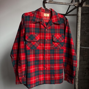 May include: Red and green plaid long-sleeve shirt with two chest pockets. The shirt has a classic collar and button-down front. The plaid pattern is a mix of red, green, and dark red. The shirt is hanging on a metal hanger.