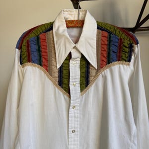 May include: White button-down shirt with a colorful, patterned yoke. The yoke features vertical stripes in red, blue, green, and tan. The shirt has a pointed collar and gold-colored buttons. The shirt is hanging on a wooden hanger.