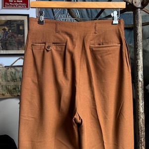 May include: High-waisted trousers in a solid rust-brown color, featuring a structured waistband with belt loops, two rear welt pockets, and a single button closure on the left pocket. The garment is suspended from a wooden hanger with metal clips.