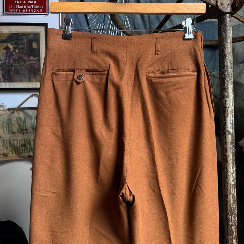 May include: High-waisted trousers in a solid rust-brown color, featuring a structured waistband with belt loops, two rear welt pockets, and a single button closure on the left pocket. The garment is suspended from a wooden hanger with metal clips.