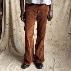 May include: A pair of brown corduroy pants with a wide-leg silhouette, featuring a button and zipper fly closure, belt loops, and a textured ribbed fabric finish, paired with black leather shoes and a plain white t-shirt.