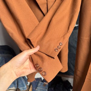 May include: A solid rust-colored blazer featuring long sleeves with three matching circular buttons on each cuff. The garment has a smooth, matte texture and a structured lapel collar, showcasing a clean and simple design throughout.