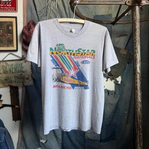 May include: A heather gray short-sleeve t-shirt featuring a graphic for the 1984 3rd Annual NorthStar Nationals NHRA drag racing event in Brainerd, Minnesota. The design includes a yellow dragster, a red star, and colorful diagonal stripes.