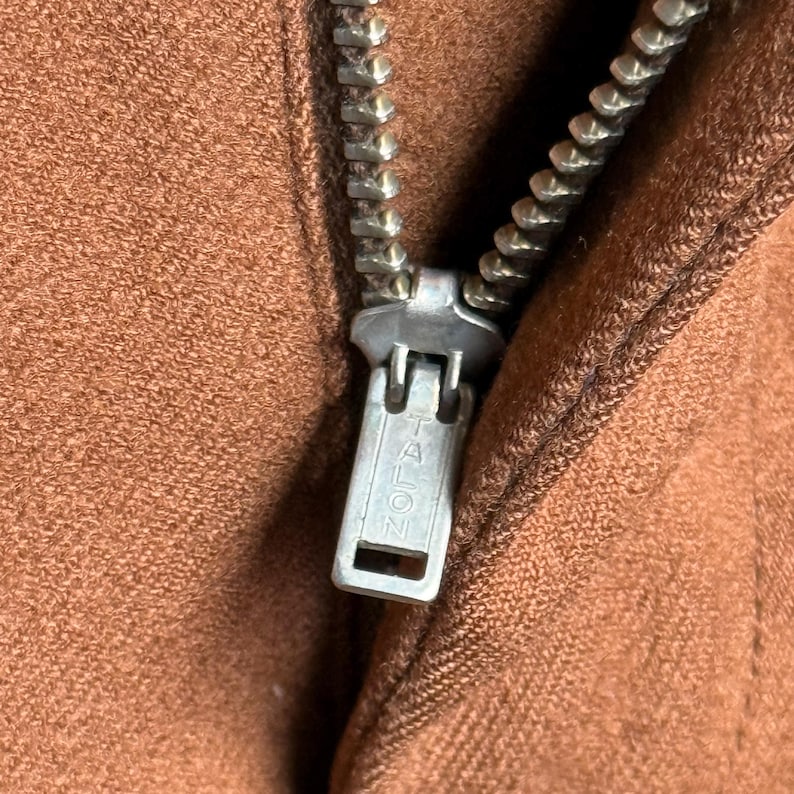 May include: A close-up view of a brown fabric garment featuring a silver-toned metal zipper. The zipper pull is clearly marked with the word TALON in embossed capital letters. The fabric has a textured, suede-like appearance with visible stitching.
