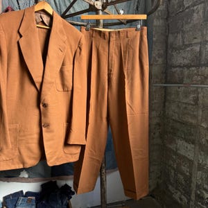 May include: A solid tan suit consisting of a two-button blazer with notched lapels and two front patch pockets, paired with matching pleated trousers featuring a cuffed hem. The set is displayed on wooden hangers.