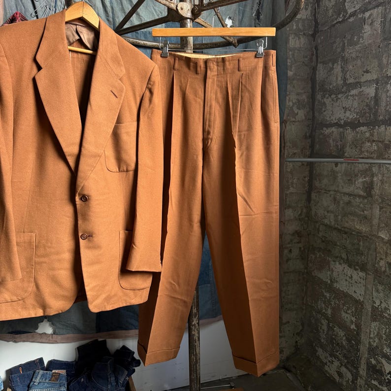 May include: A solid tan suit consisting of a two-button blazer with notched lapels and two front patch pockets, paired with matching pleated trousers featuring a cuffed hem. The set is displayed on wooden hangers.