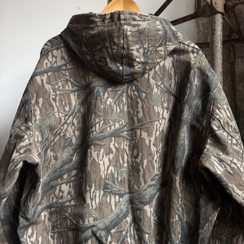 Puede incluir: Una sudadera con capucha con un patr&oacute;n de camuflaje. El patr&oacute;n presenta una mezcla de tonos marrones, verdes y grises, que se asemejan a una escena forestal. La sudadera con capucha est&aacute; colgada de una percha de madera.