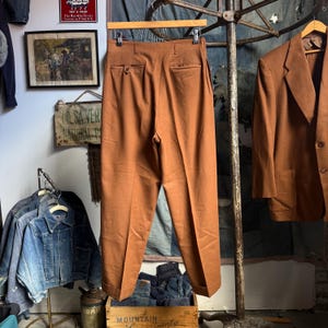 May include: A pair of high-waisted trousers in a solid rust-brown color, featuring a straight-leg cut with cuffed hems, a buttoned rear pocket, and a clean, tailored finish. The trousers are suspended from a wooden hanger.
