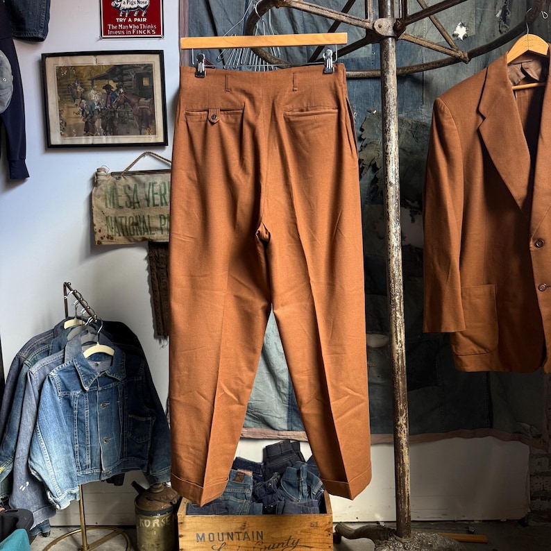 May include: A pair of high-waisted trousers in a solid rust-brown color, featuring a straight-leg cut with cuffed hems, a buttoned rear pocket, and a clean, tailored finish. The trousers are suspended from a wooden hanger.
