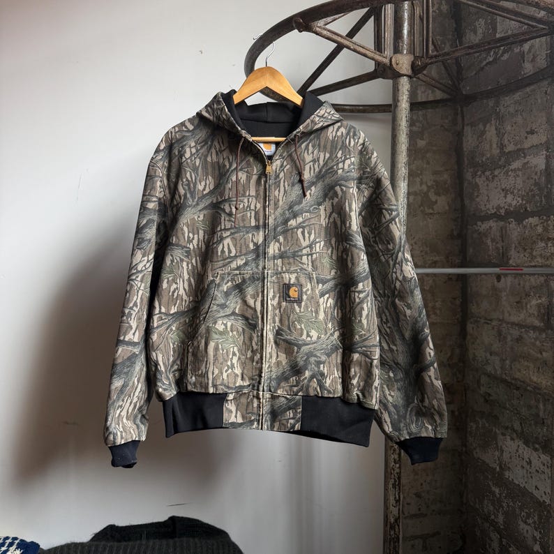 Puede incluir: Chaqueta con capucha y cremallera con estampado de camuflaje en tonos marrones, grises y verdes. La chaqueta tiene forro, pu&ntilde;os y dobladillo negros. Un peque&ntilde;o logotipo cuadrado es visible en la parte delantera.