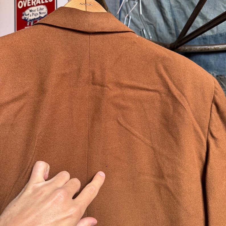 May include: A solid brown blazer featuring a notched lapel collar and a central vertical seam down the back. The fabric has a soft, matte texture. A small, dark spot is visible on the lower center of the back panel of the garment.
