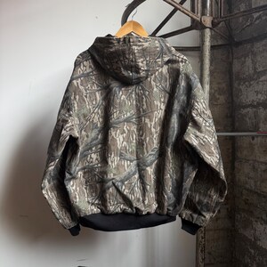 Puede incluir: Chaqueta con capucha de camuflaje en tonos marrones, verdes y grises. La chaqueta tiene un dobladillo y pu&ntilde;os de canal&eacute; negros. La parte trasera de la chaqueta es visible, colgada de una percha de madera.