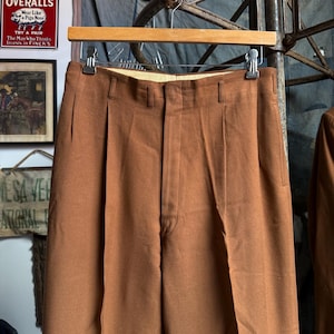 May include: A pair of solid brown trousers featuring a high-waisted design with double front pleats, a central zipper fly, and belt loops. The trousers are suspended from a wooden hanger with metal clips, showcasing a smooth, matte fabric finish.