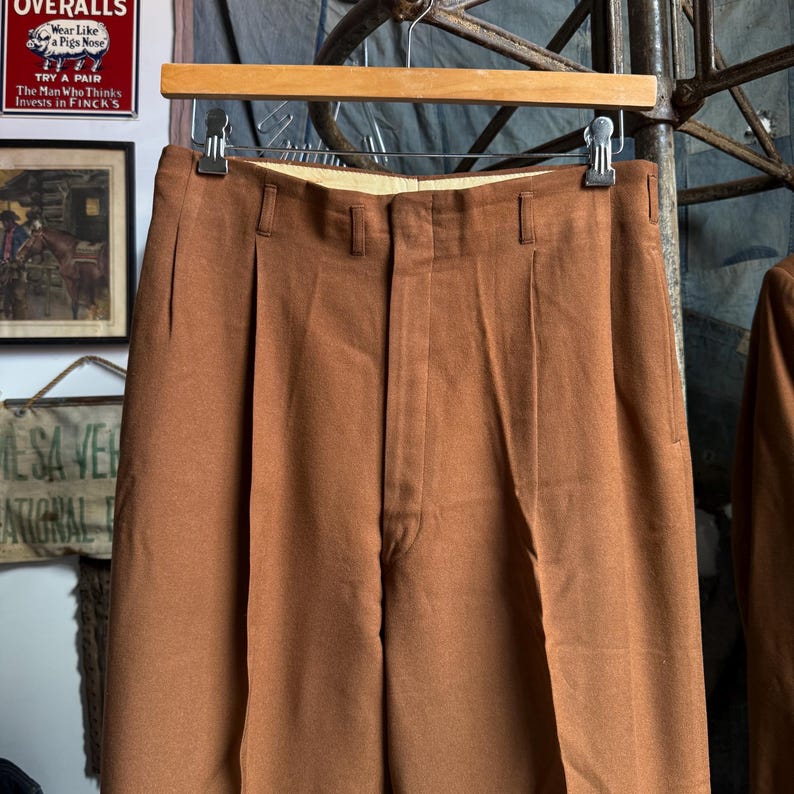May include: A pair of solid brown trousers featuring a high-waisted design with double front pleats, a central zipper fly, and belt loops. The trousers are suspended from a wooden hanger with metal clips, showcasing a smooth, matte fabric finish.