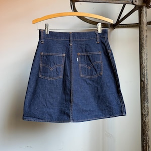 May include: A dark blue denim skirt with two back pockets and orange stitching. The skirt is hanging on a wooden hanger. The Levi's brand label is visible on the back pocket.