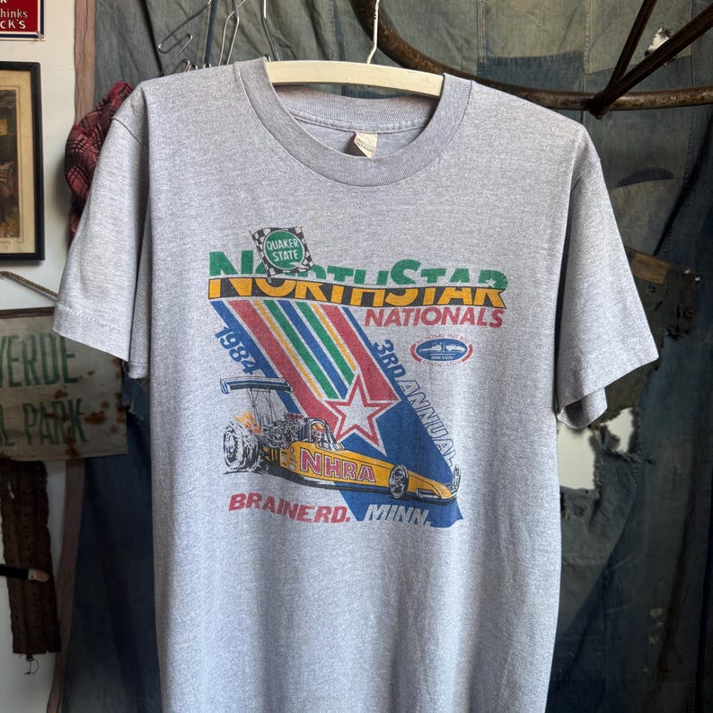 May include: A heather gray short-sleeve t-shirt featuring a graphic for the 1984 3rd Annual North Star Nationals NHRA drag racing event in Brainerd, Minnesota. The design includes a yellow dragster, a red star, and Quaker State branding.