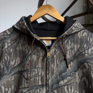 Puede incluir: Chaqueta con capucha Carhartt con un estampado de camuflaje en marr&oacute;n, gris y verde. La chaqueta tiene una cremallera completa, un forro de capucha negro y cordones marrones. El logotipo de Carhartt es visible en el pecho.