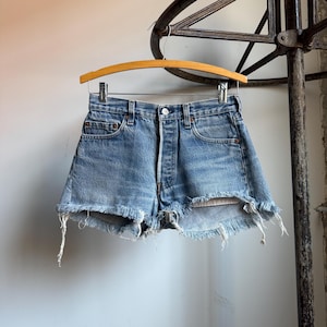 May include: Light blue denim shorts with a frayed hem, hanging on a wooden hanger. The shorts have a button fly and classic five-pocket design. The denim is faded, giving a vintage look.