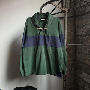 May include: Green long-sleeve shirt with a navy blue horizontal stripe across the chest. The shirt has a collared neckline with a wooden toggle closure. The brand name "Orvis" is visible on the collar.